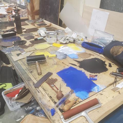 Photo du plan de travail d'impression des linogravures : rouleaux, encres étalées sur une plaque de verre, plaques de lino, beaucoup de bordel.
