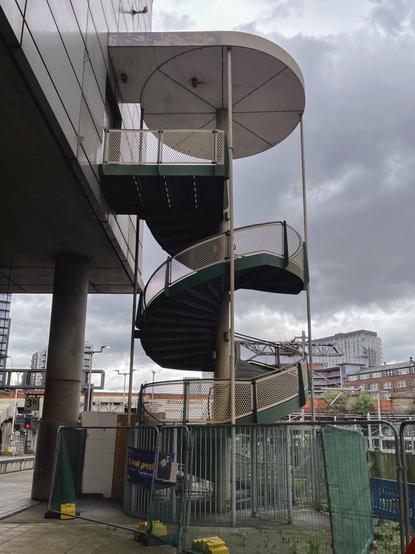 A green metal spiral staircase with mesh railings stands outdoors, sheltered partially by round roofs. It is attached to a modern building, next to a fence and construction barriers, with a cloudy sky and city buildings in the background.