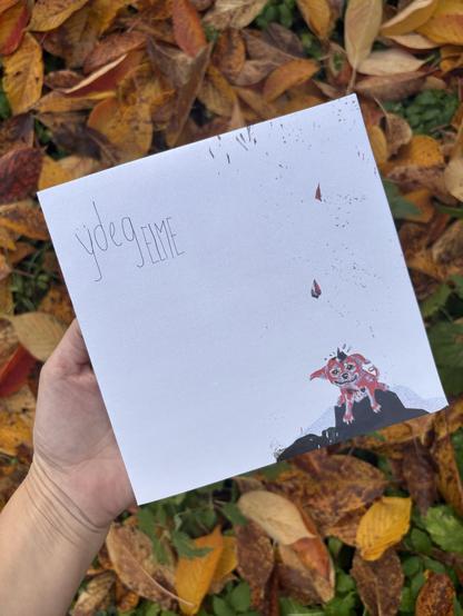 Eine Hand hält ein weißes Blatt Papier mit dem Text „Ÿdeg ELME” und einer Illustration eines kleinen Hundes auf einer felsigen Oberfläche. Der Hintergrund besteht aus bunten Herbstblättern.