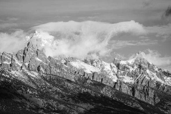 The snow-covered jagged peaks of the Cathedral Group of the Teton Range, with low clouds partially obscuring Grand Teton and forested lower slopes leading toward the foreground.