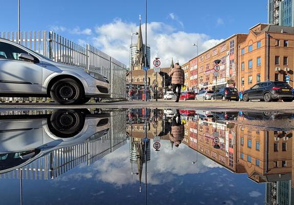 A city street scene with buildings, cars, and a pedestrian reflected perfectly in a large puddle, creating a symmetrical mirrored effect.