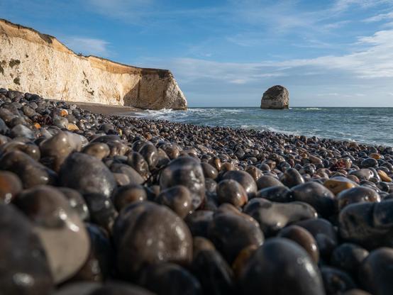 A low-angle view across smooth dark pebbles toward white coastal cliffs and a single offshore rock beneath a blue sky.