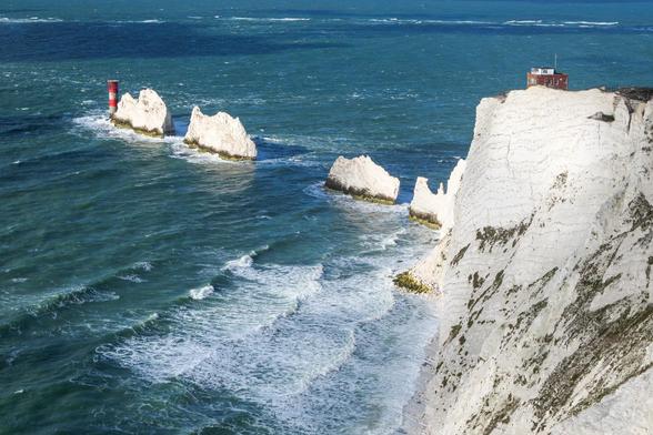 A high viewpoint looking down on turquoise sea, white chalk cliffs, and a series of rock stacks with a small red-and-white lighthouse.