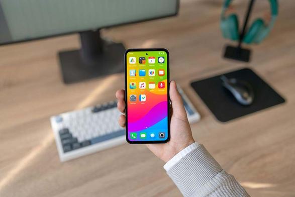 Hand holding the Murena Fairphone (Gen. 6) running /e/OS and a keyboard, a mouse and a screen on a desktop as background