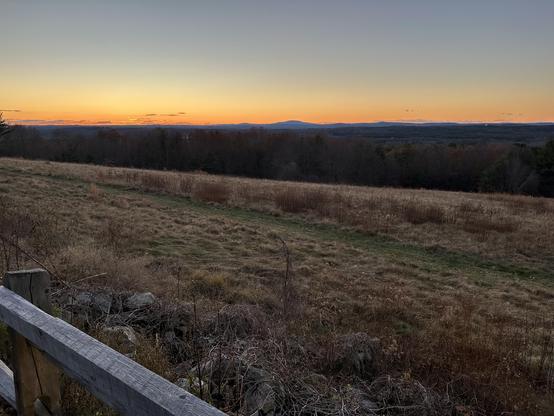 A wide, open landscape at sunset with a glowing orange horizon fading into a clear blue sky. Rolling hills and distant mountains form a dark silhouette beneath the setting sun. In the foreground, there is a grassy, slightly uneven field with patches of dry vegetation and a low wooden fence running along the left edge. Beyond the field, leafless and evergreen trees create a dark band of forest leading toward the distant hills. The scene feels calm, quiet, and cold, with the last light of day illuminating the sky.