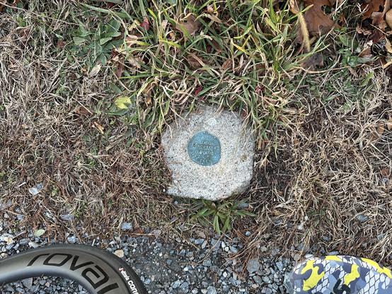 A small round metal survey marker is embedded in a rough concrete block set into the ground, surrounded by dry grass and a few green plants. The marker has engraved text, though it’s partially worn and difficult to read. At the bottom left edge of the frame is the rim of a bicycle wheel with the “Roval” logo, and at the bottom right edge is part of a shoe with a yellow, gray, and black camo pattern.