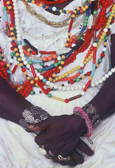 Close-up of a person wearing numerous colorful beaded necklaces and heavy silver and beaded bracelets over a white garment, with hands clasped and adorned with several rings.