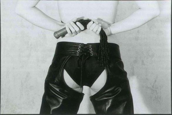 Black and white close-up of a person wearing high-waisted, lace-up leather chaps while holding the handle of a braided whip behind their back.