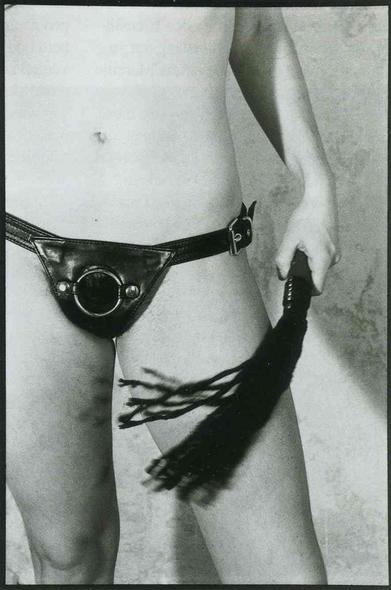Black and white close-up of a person wearing a leather harness with a metal ring, holding a black tassel whip.