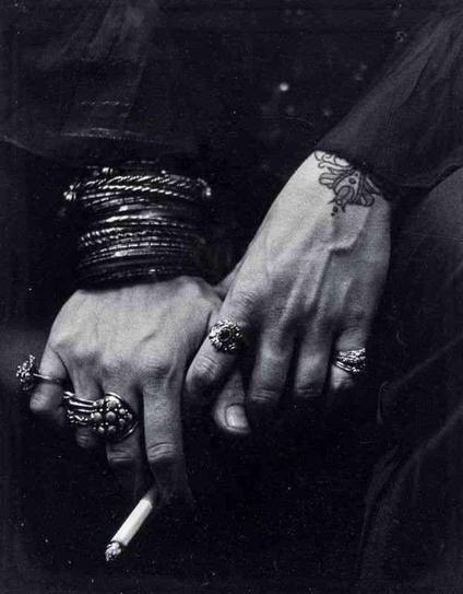 Close-up black and white photograph of two hands adorned with multiple ornate rings and bracelets, one hand holding a cigarette.