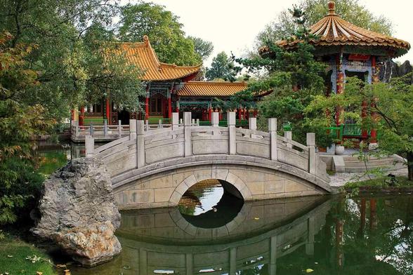 Arched stone bridge crossing green water in a lush Chinese garden featuring traditional pavilions with orange tiled roofs.