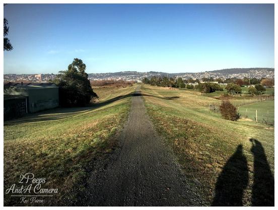 A wide, coloured landscape photograph showing a long, straight gravel path receding directly into the distance between two gently sloping grassy embankments.

The path appears to be on an elevated strip of land. In the background, the houses of Launceston stretch across the low hills under a bright blue sky. A large shadow is cast in the lower right corner.