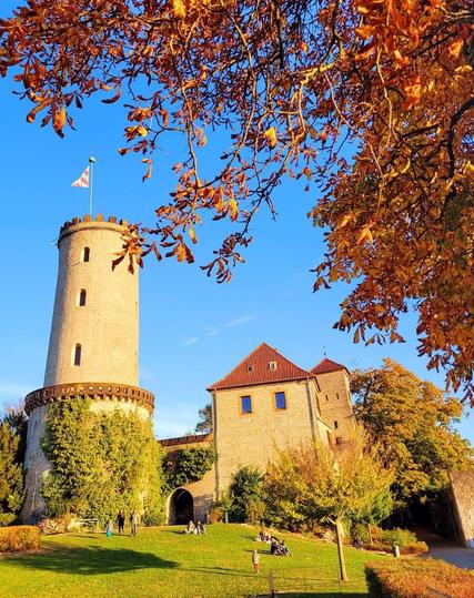 Herbstliche Szene einer Burganlage in Bielefeld: Ein hoher runder Steinturm mit wehender Flagge und ein angrenzendes Gebäude mit rotem Dach stehen vor blauem Himmel. Im Vordergrund sind goldene und orangefarbene Herbstblätter an den Ästen eines Baumes zu sehen. Menschen sitzen und spazieren auf einer sonnigen Rasenfläche am Fuß der Burg.