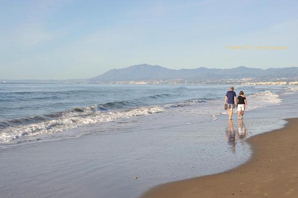 Imagen de una playa donde se ven dos personas paseando por la orilla del mar. A lo lejos se ve una montaña. Cielo y mar ocupan la mayor parte del cuadro.