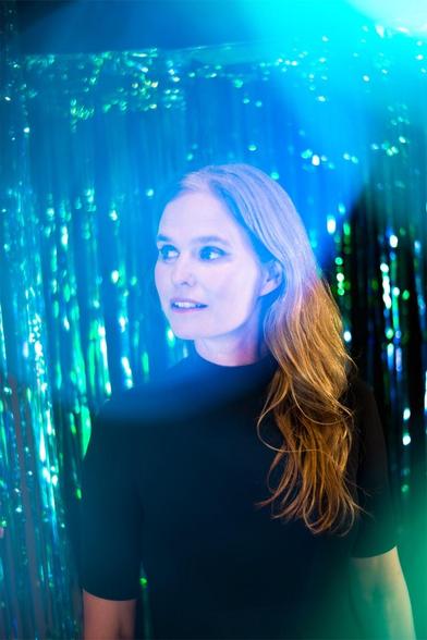 Eine Frau mit langen, welligen Haaren blickt vor einem Hintergrund aus schimmernden blauen und grünen Lichtern zur Seite. Sie trägt ein schwarzes Oberteil und hat einen nachdenklichen Gesichtsausdruck. Die Beleuchtung schafft eine lebendige und dynamische Atmosphäre.