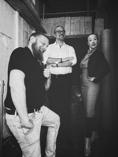 S/W-Bild von zwei Männern und einer Frau in Kostümen, die Grimassen schneidend in einem Hinterbühnenbereich stehen. 