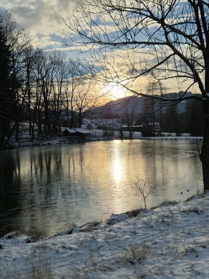 A serene winter landscape unfolds as the sun begins to set, casting a warm, golden glow across a tranquil river. The river, partially covered in a thin layer of ice, reflects the soft light of the setting sun and the bare branches of the surrounding trees. The trees, stripped of their leaves, stand tall and stark against the pale blue sky, their silhouettes adding a sense of calm and stillness to the scene.

The riverbank is dusted with a light layer of snow, creating a delicate contrast with t…