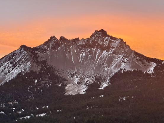 A rugged, snow-dusted mountain rises above a dense evergreen forest at sunrise. The sky glows with vivid orange and pink tones, casting warm light on the jagged peaks. Snow highlights steep chutes and rocky textures across the mountain’s face.