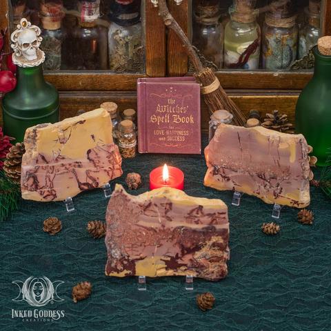 Three specimens of the "Mookaite Jasper Altar Tile" product are propped up on a lace-covered surface. The flat, roughly rectangular slices are about the size of a bar of soap and feature layered patterns of yellow and pink with brown veins. Presented by Inked Goddess Creations.