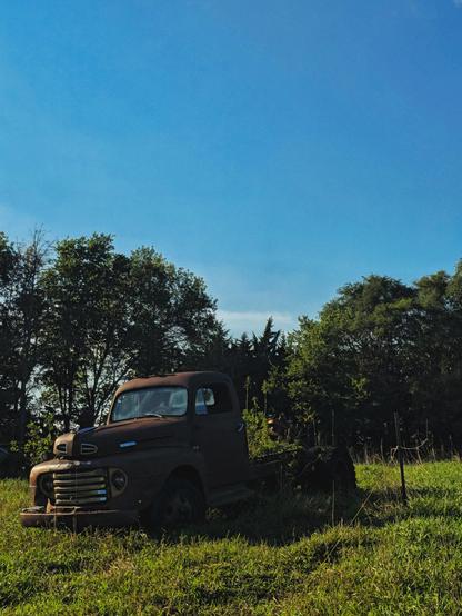 An old rusted truck rests in a sunny field with tall grass and trees in the background against a clear blue sky, conveying a sense of nostalgia and tranquility.