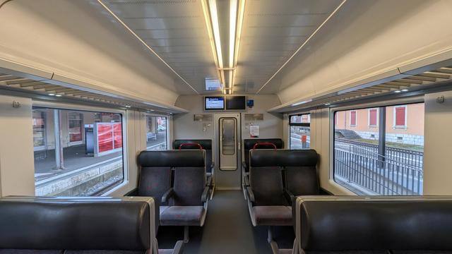 Interior of the CJ class RBDe 567 motor coach, dark-gray vis-à-vis seat benches with foldable armrest in the middle, white wall and ceiling panels, grey-silver partition to the vestibule, two screens above the door, left one showing the stops