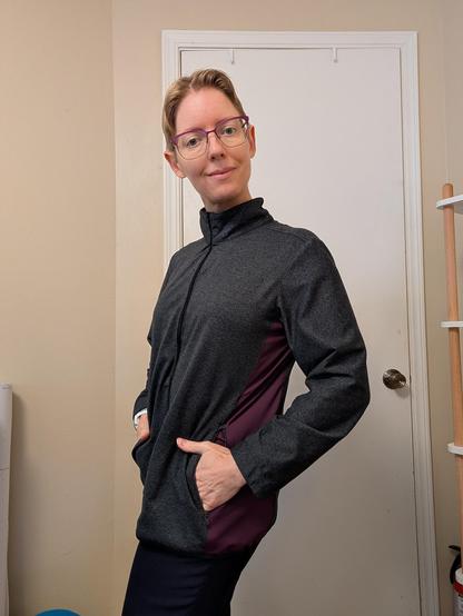 Blonde woman in a charcoal grey jacket with a plum side panel.  Her hands are in zippered side pockets and the jacket is zipped all the way up.