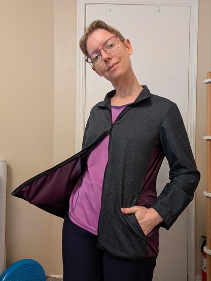 Blonde woman in a charcoal grey jacket with a plum side panel. The jacket is unzipped except for a small portion near the top, the collar is folded down, and one hand is in a side pocket.  The other hand is holding the jacket open, showing an interior pocket that is about half as long as the jacket itself!