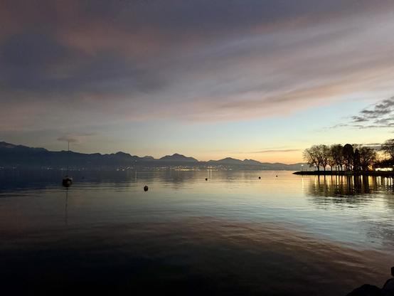 Lac Leman at dusk. Low orange glow on the horizon, turquoise sky with billowing grey clouds relecting pink and orange highlights. A line of trees, bare of leaves, in silhouette. Distant mountains. All reflected in the dark undulating surface of the water.