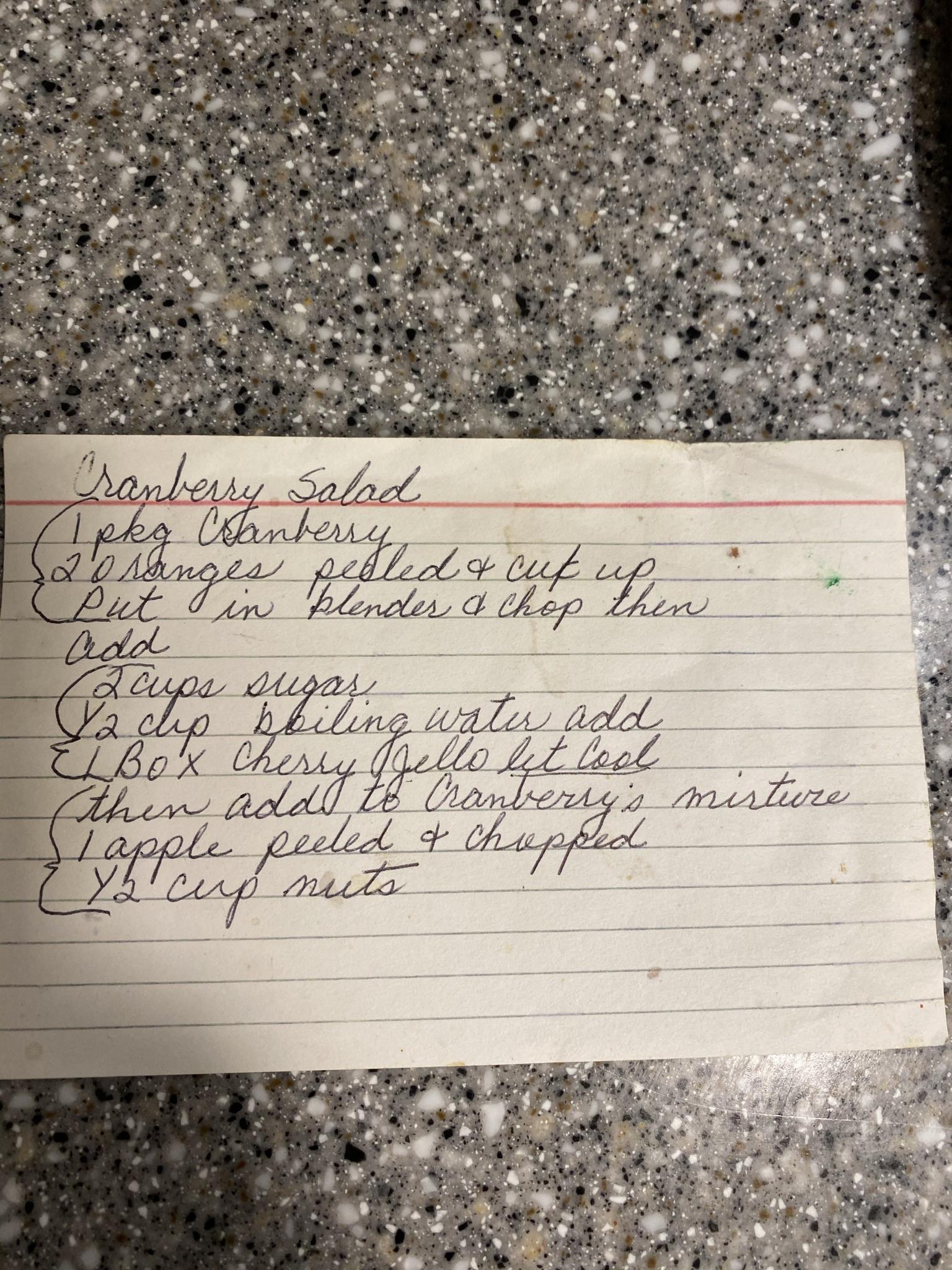 A handwritten recipe card for "Cranberry Salad," listing ingredients and preparation steps, including cranberries, oranges, sugar, cherry Jell-O, apple, and nuts. The card is placed on a speckled countertop.