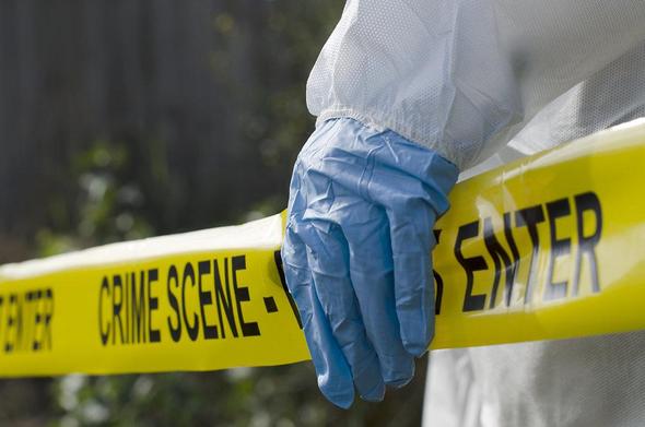 Hand in blue glove holding yellow tape reading "CRIME SCENE." Background is blurred, creating a tense, investigative mood.