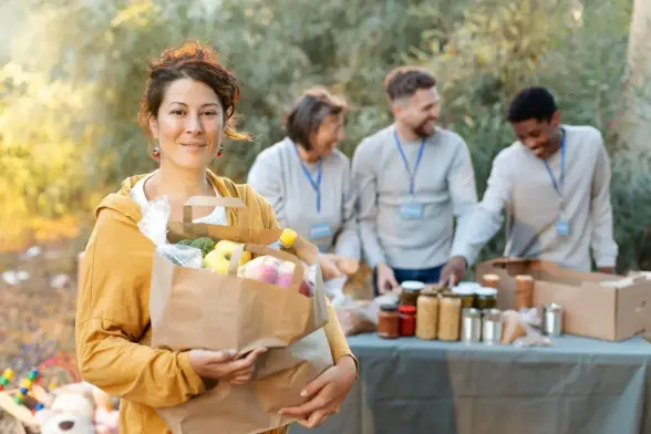 A smiling woman with reddish brown hair tied up stands in the foreground, holding a full paper grocery bag containing fresh vegetables (broccoli, apples, bananas). She wears a mustard yellow hoodie and large earrings. Behind her, slightly out of focus, three volunteers in gray shirts and blue lanyards sort canned goods and jars on a folding table at an outdoor food drive. Autumn trees and soft sunlight create a warm, community atmosphere.