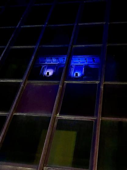 Reflejo de unas ventanas y algunas molduras, de color morado por la luz nocturna, en una fachada de cristal. El mismo lugar fotografiado de dos maneras diferentes, una con más luz y otra más negra.