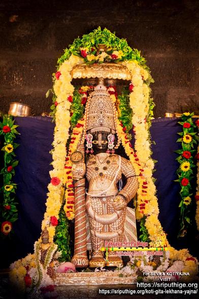 Idol of Hindu deity Krishna in diamond cladding and a matching crown, holding a similarly clad staff in right hand, a small gold umbrella overhead. A string of alternating yellow and red flowers around Krishna waist to waist. A thick string of yellow flowers around Krishna floor-to-floor going over the umbrella. A string of green holy basil leaves atop the umbrella. Dark blue cloth background. A string of green betel leaves (alternating red and yellow flowers on the leaves) hangs down at each end of the background.