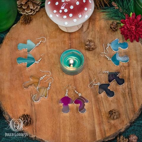 Five example pairs of the "Dyed Agate Mushroom Earrings" product are displayed a round wood slice. The agates are carved into small mushroom shapes and range in colors of pink, gold, black, teal, and blue. They have silver solder around the outside of the carving and hang from silver fishhook earwires. Presented by Inked Goddess Creations.