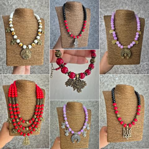 A collage of seven photographs depicting handmade necklaces in the traditiona Ukrainian style. The necklaces are made of large colored beads (red, black, white, lilac) and decorated with metal pendants: crosses, images of saints. the Trident (the coat of arms of Ukraine), and symbolic crescent-shaped pendants. The necklaces are displayed on mannequins covered in burlap.