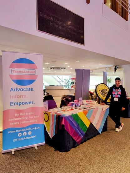 Image shows one of our directors, Maryam, standing next to a table which has  different pride flags on it with our leaflets and reports. Next to the table is a standing banner with the TransActual logo and the words "Advocate. Empower. Inform. By the trans community, for the trans community."
