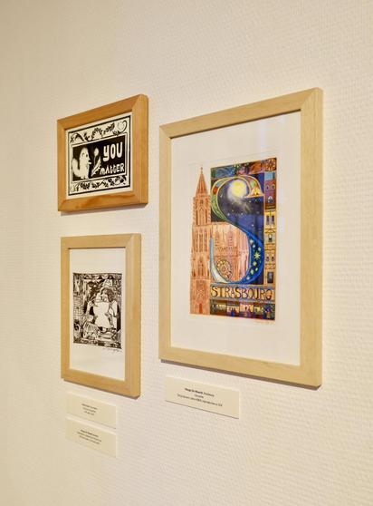 Trois œuvres de l'exposition : en haut à gauche, une linogravure intitulée « You matter » (Tu comptes) de Daisy Gand. Les deux autres œuvres sont de moi : celle de gauche est un dessin à l'encre noir et blanc intitulé « Recette » au format A5. Celle de droite est une peinture à la gouache au format A5 dans un cadre A4, intitulée « Strasbourg ».