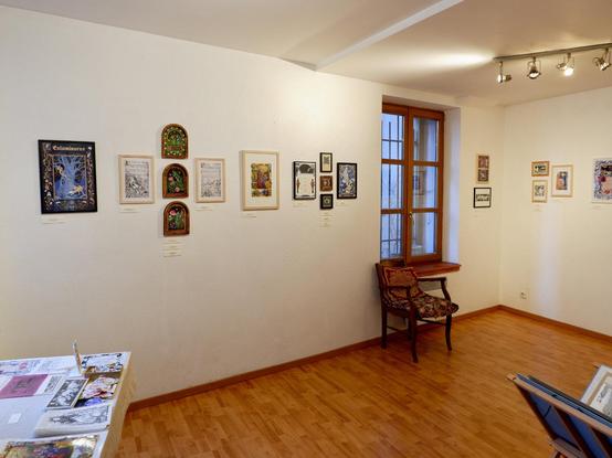 Une photo de l'espace d'exposition où l'on voit les peintures et les œuvres accrochées au mur. Sur les bords, on aperçoit une table avec des tirages et des articles dérivés, ainsi qu'un dossier contenant des tirages grand format.