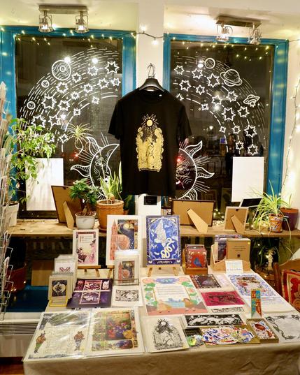 Une des tables avec des gravures et des articles dérivés. Elle se trouve devant deux fenêtres décorées de dessins représentant la lune, le soleil et les étoiles, et les cadres des fenêtres sont ornés de guirlandes lumineuses.