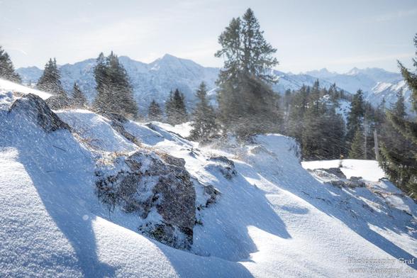 A breathtaking winter landscape unfolds under a clear, bright sky, where the sun casts a brilliant light over snow-covered mountains and rocky terrain. The scene captures a moment of fresh snowfall, with flurries gently drifting through the air, adding a sense of movement and tranquility. The snow blankets the jagged rocks and uneven ground, creating a striking contrast between the white snow and the darker, rugged surfaces beneath.

In the background, a dense forest of evergreen trees stretche…