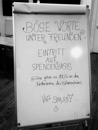 S/W-Bild eines aufgestellten Whiteboards, auf dem in schnörkeliger schwarzer Schrift „BÖSE WORTE UNTER FREUNDEN - EINTRITT AUF SPENDENBASIS - Erlöse gehen zu 100% an den Förderverein des Kulturzentrums - VIEL SPASS! :-)“ steht. 