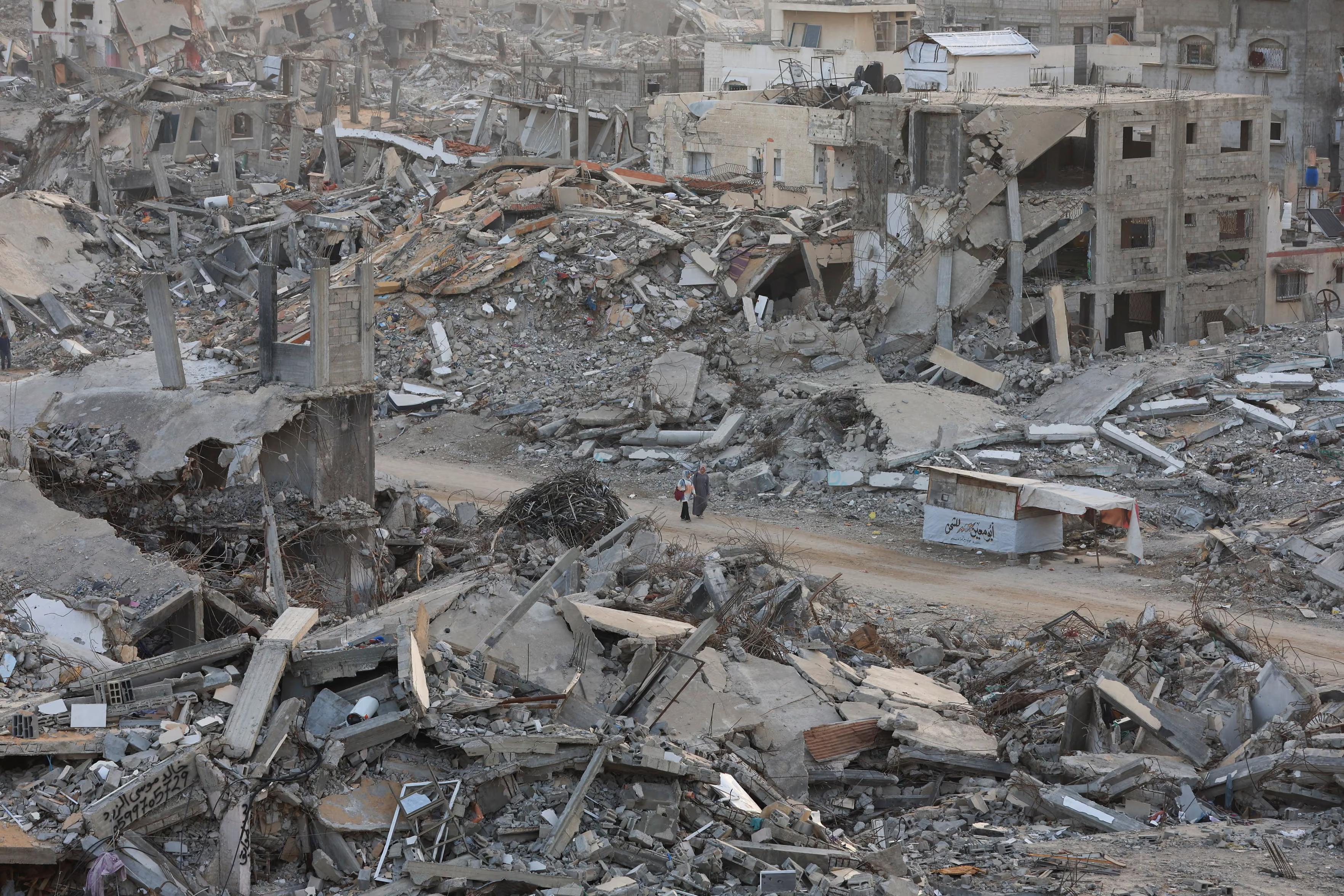Two Palestinian women walk among mounds of rubble and destroyed buildings.