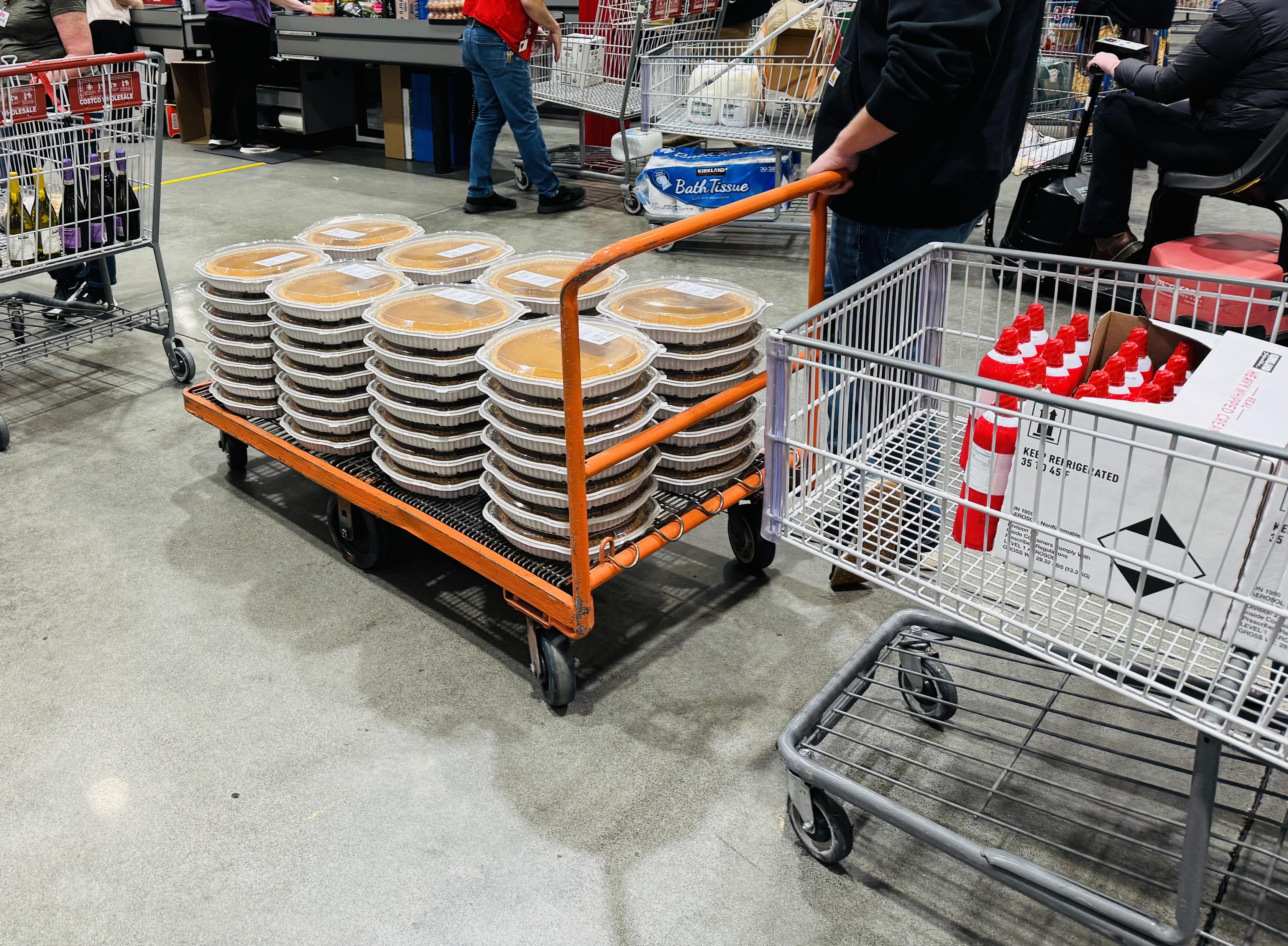 About 60 oversized pumpkin pies and a similar number of spray whipped cream cans on a flat bed cart at Costco checkout.