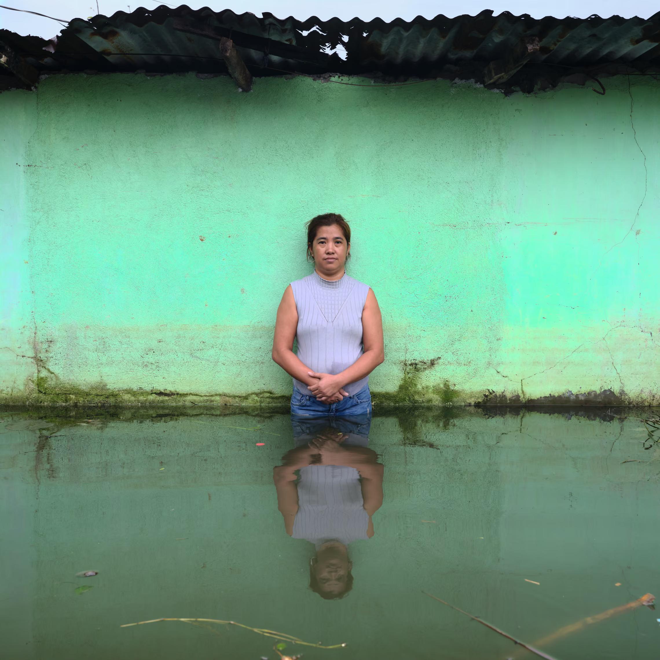A woman standing in water outside her house, her image reflected in the water.
"...they shouldn’t steal and engage in corrupt practices.They shouldn’t be stealing from people; they should be helping their constituents so that our town won’t be submerged by the floodwaters."