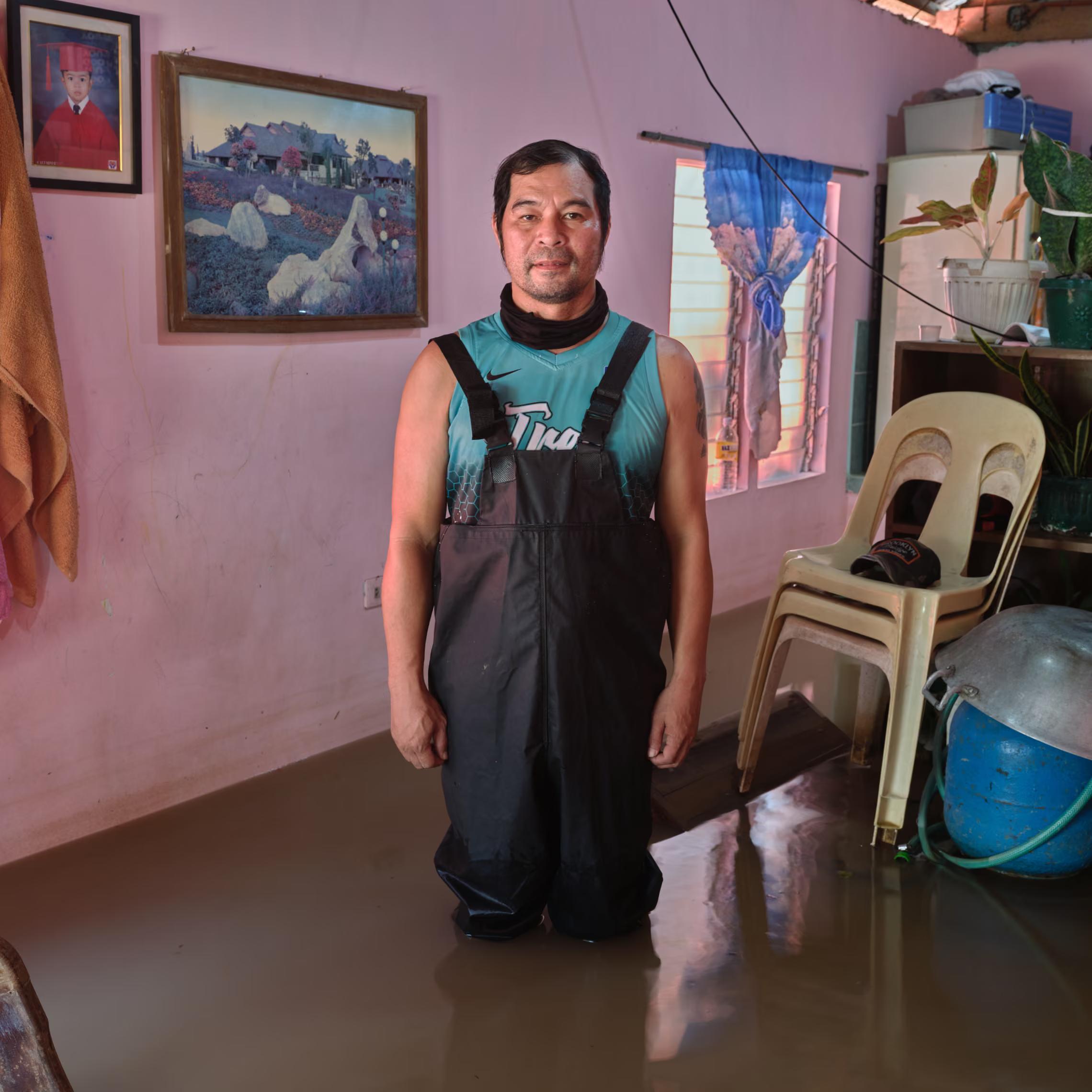 A man in waders stands inside his flooded room with pictures on the walls.
"Due to climate change, the flooding is getting worse. Ten years ago, the roads were dry … we didn’t have to elevate our houses. I am afraid for our children, for everybody … What will happen if we don’t find a solution to this?"