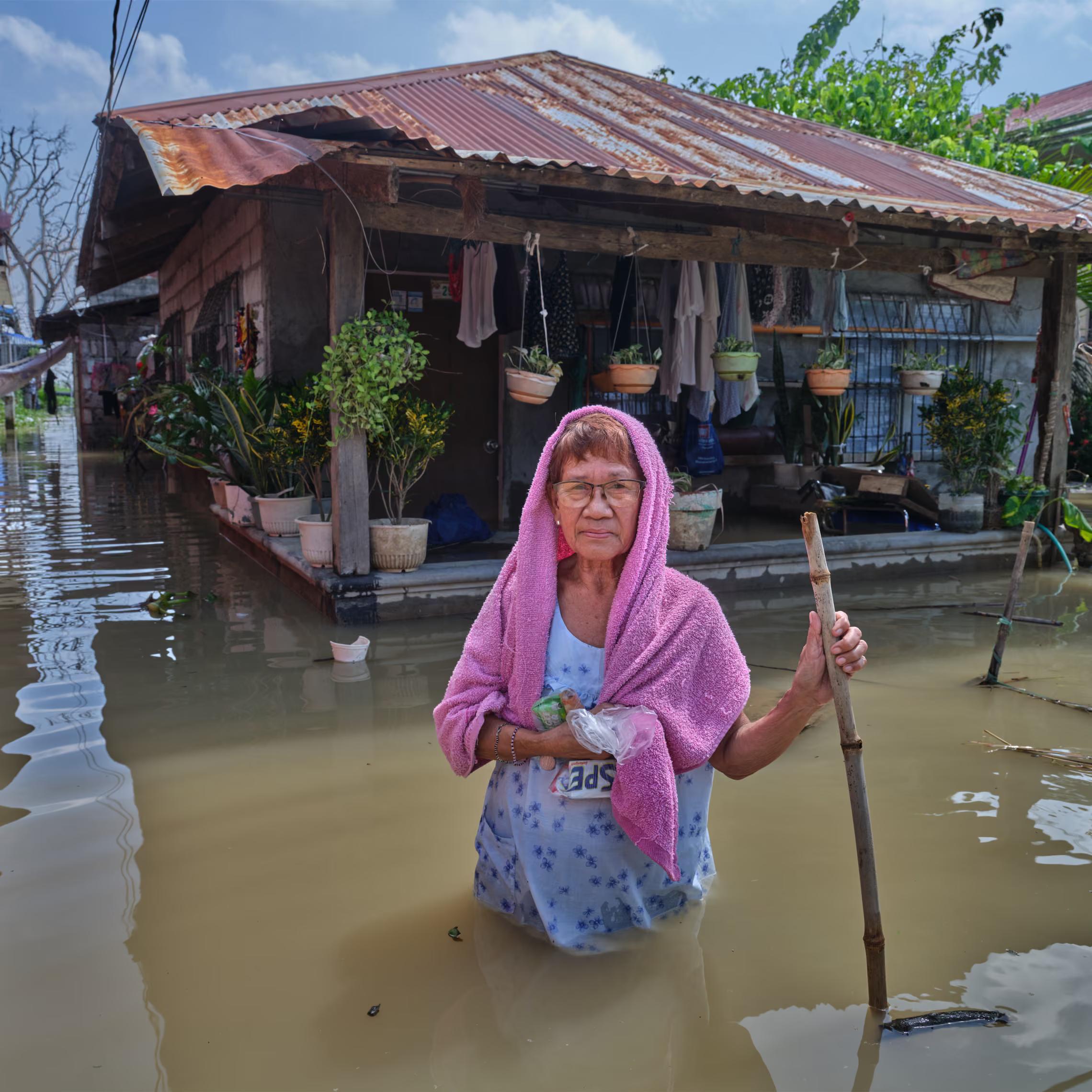 A woman with a pink shawl stands outside her house in raised waters, with pot plants on the porch.
"The water is rising. Because of the frequent rain and storms, right? We’re behind that river, so all the water from the country passes here on the way to the sea. We’re really used to that water every year."