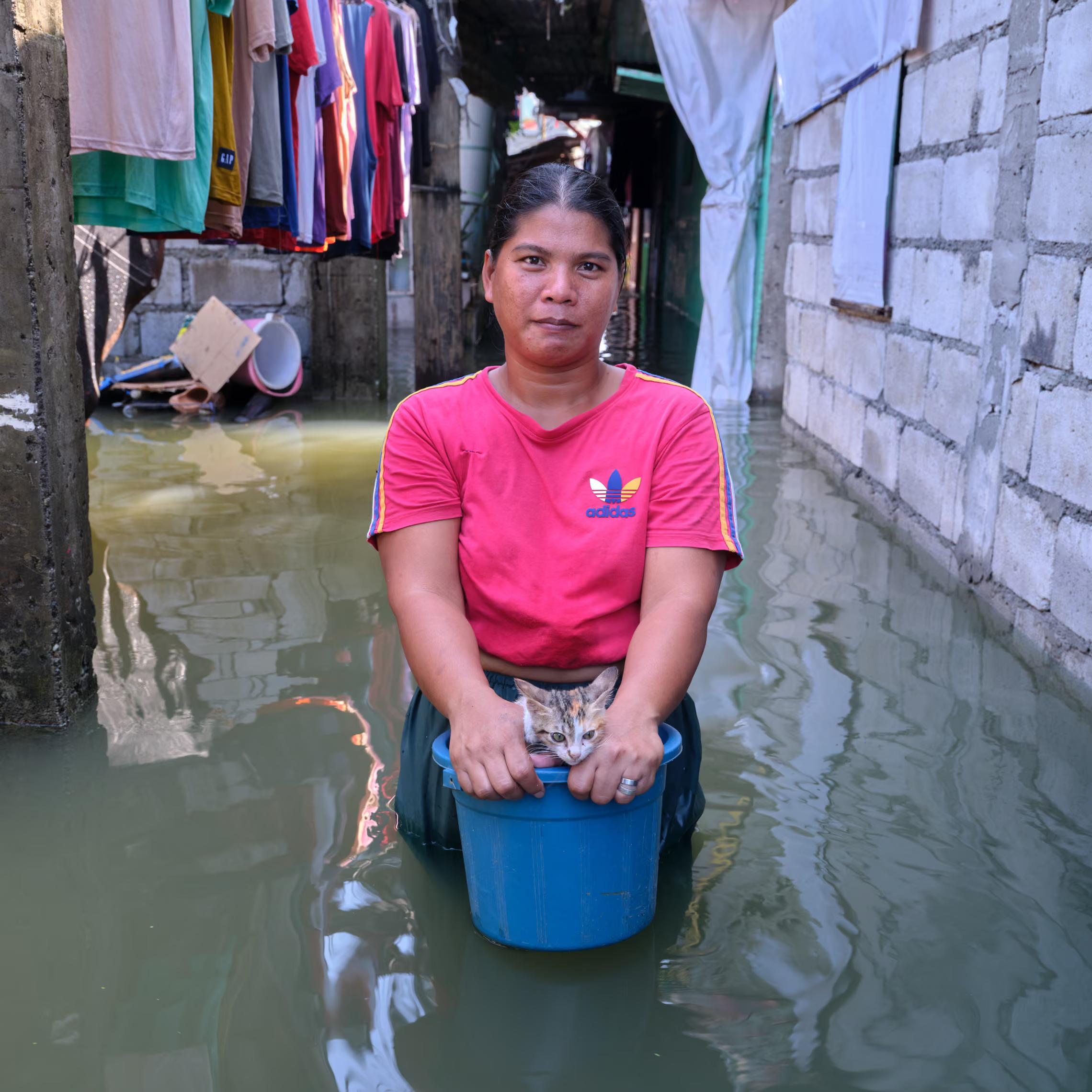 A woman stands in water holding a bucket. There's a rack of clothes hanging high behind her.
"Then the storm came along, and the dams were released at the same time. That’s why the water got deeper. I hope the flood and high tide won’t occur again so children will be able to play in the street and can have more fun."