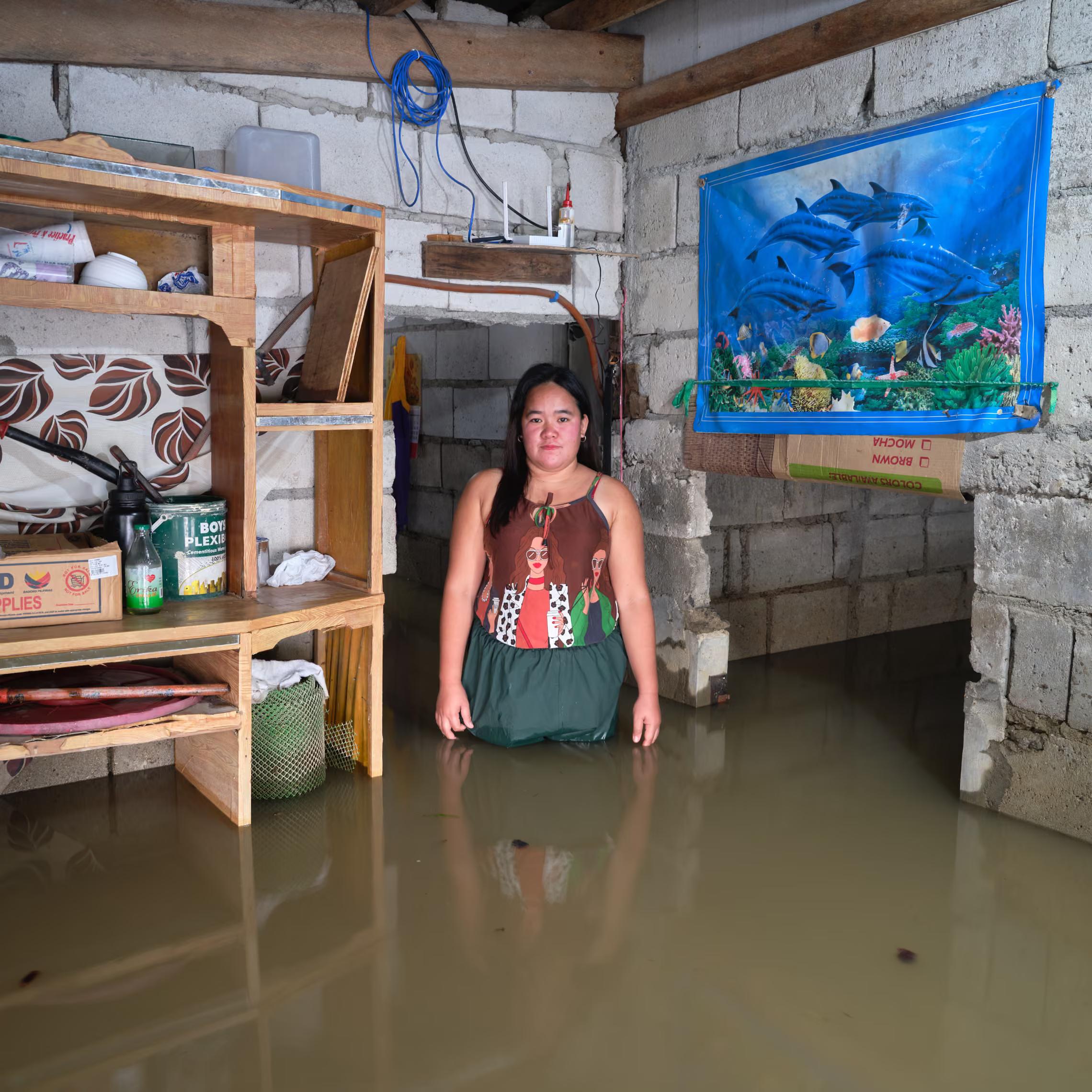A young woman stands in water inside her house with a picture of dolphins on the wall.
" I hope a solution can be found so that we will never experience this suffering again. I hope the government budget will be used properly so that something can be done about our situation."