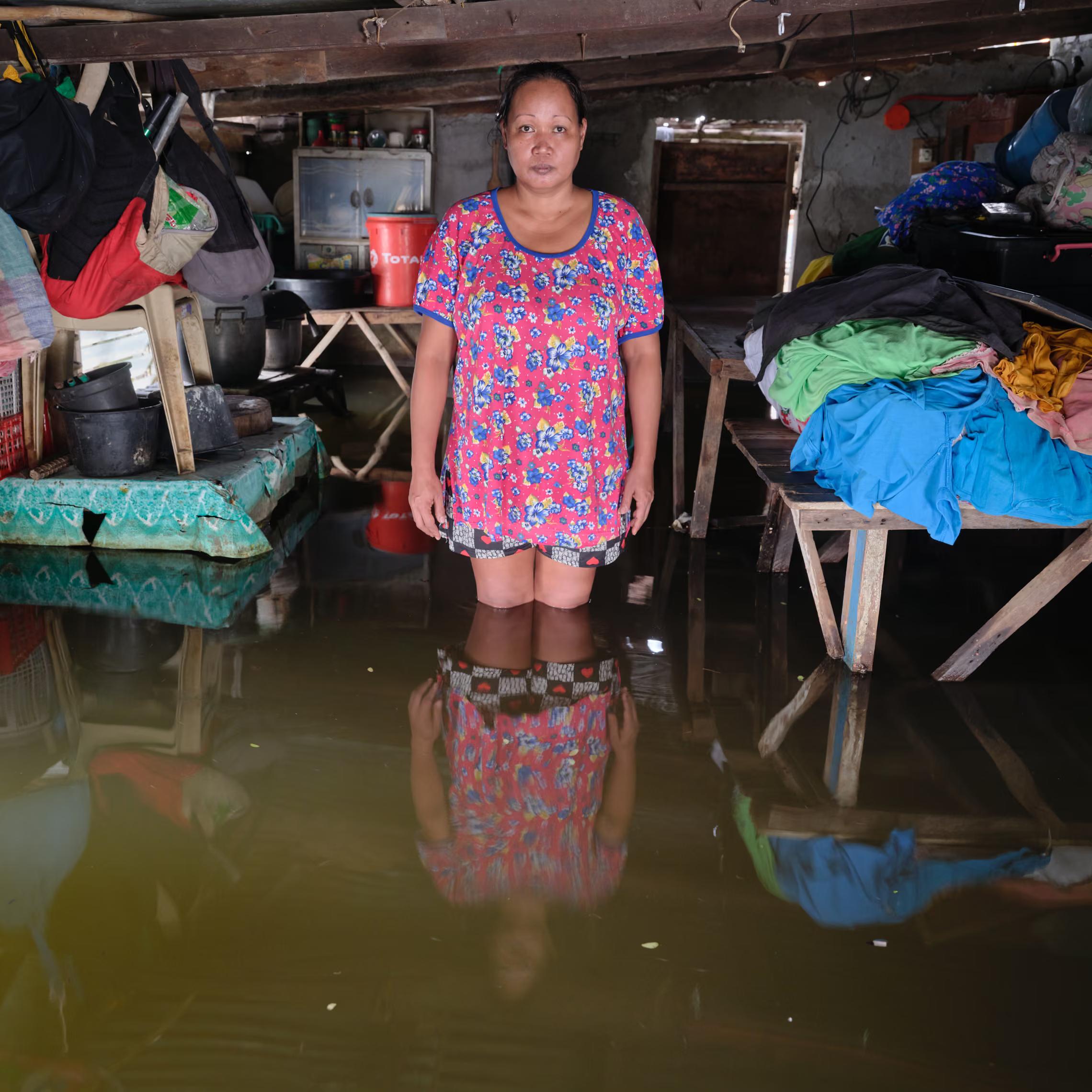 A woman in a pink dress stands i a knee-deep water in her flooded house, with their belongings piled up high.
" I hope we can get help with the problem of rising water, which has been here since Typhoon Emong. The water has been increasing; it’s not disappearing any more. That’s the change I’ve seen here."