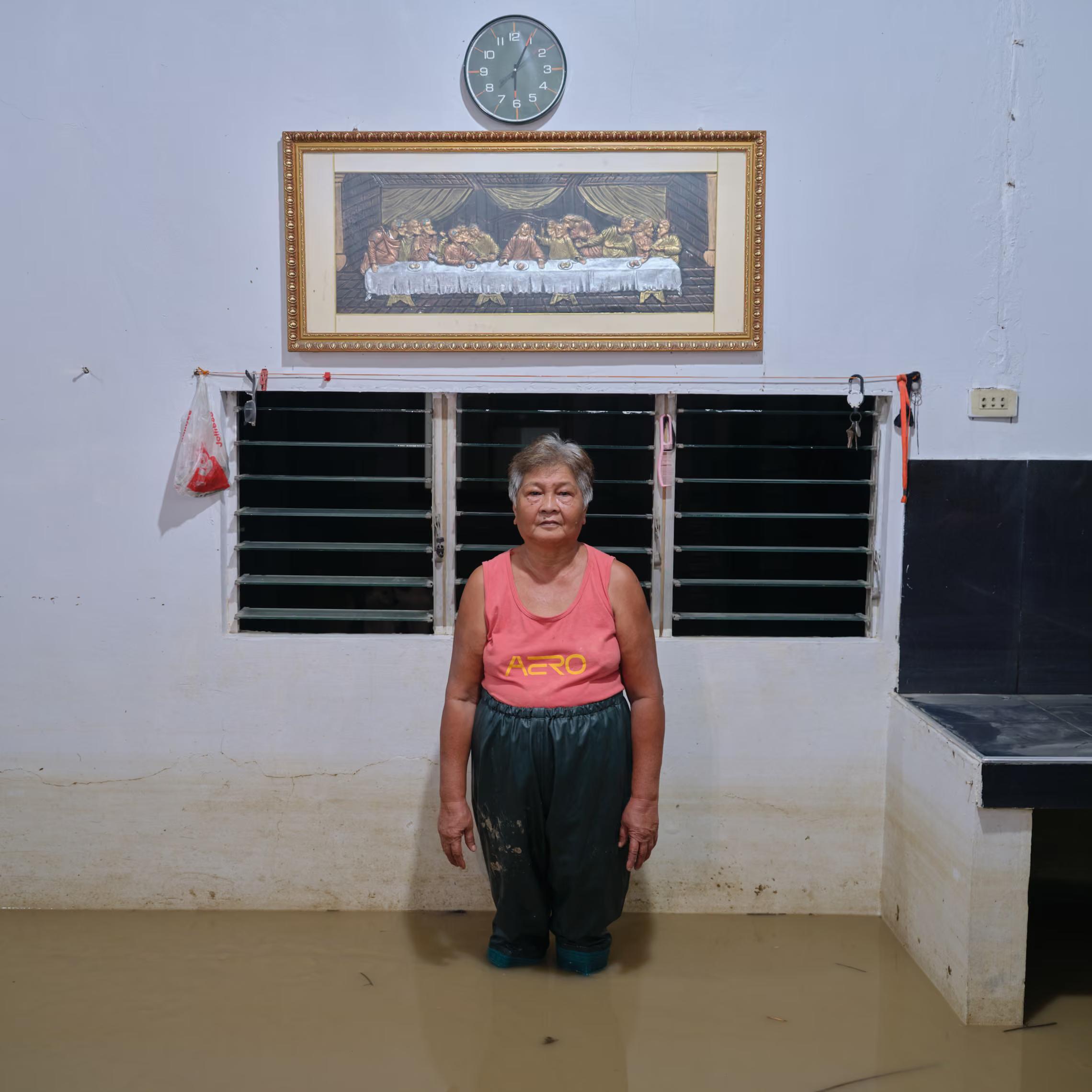 A 65-year-old woman stands in her flooded room, the picture of The Last Supper behind her.
"But when it floods now, it goes up and down until we’ve reached seven months of mud and water. They shouldn’t cut down the trees on the mountains and block water channels that let the water flow straight out."